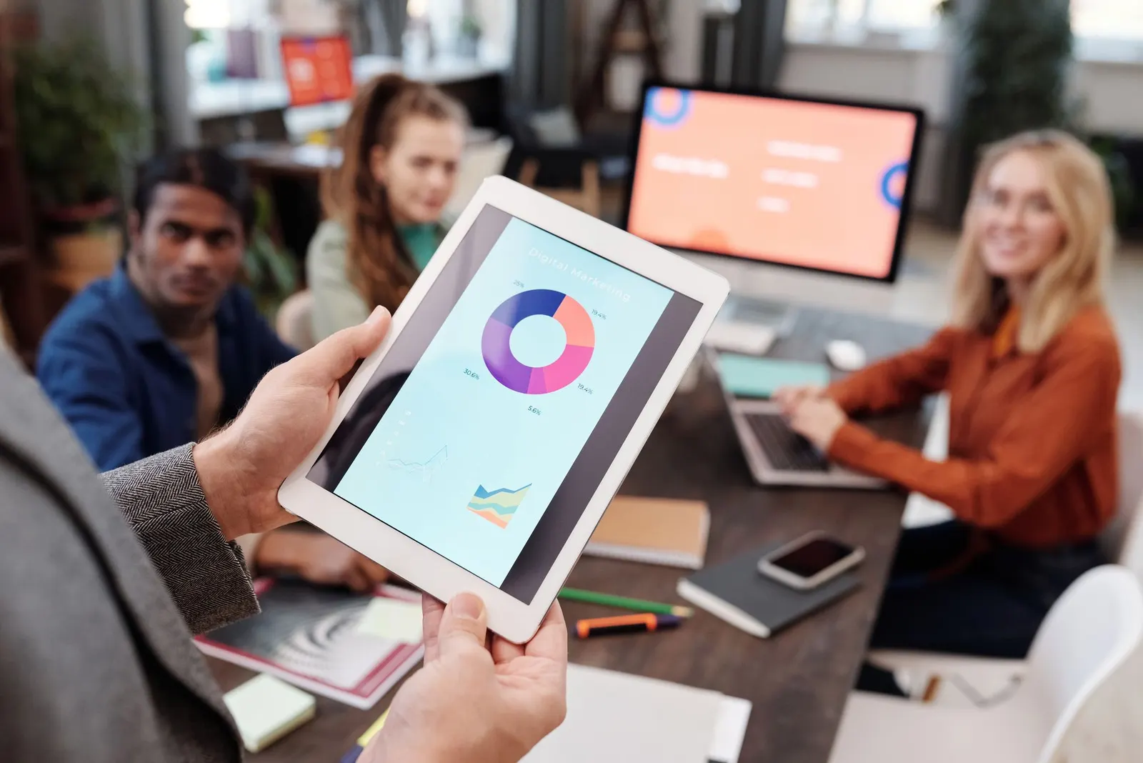 Person hält ein Tablet mit einem Kreisdiagramm, im Hintergrund ein Team bei der Auswertung einer Kampagne