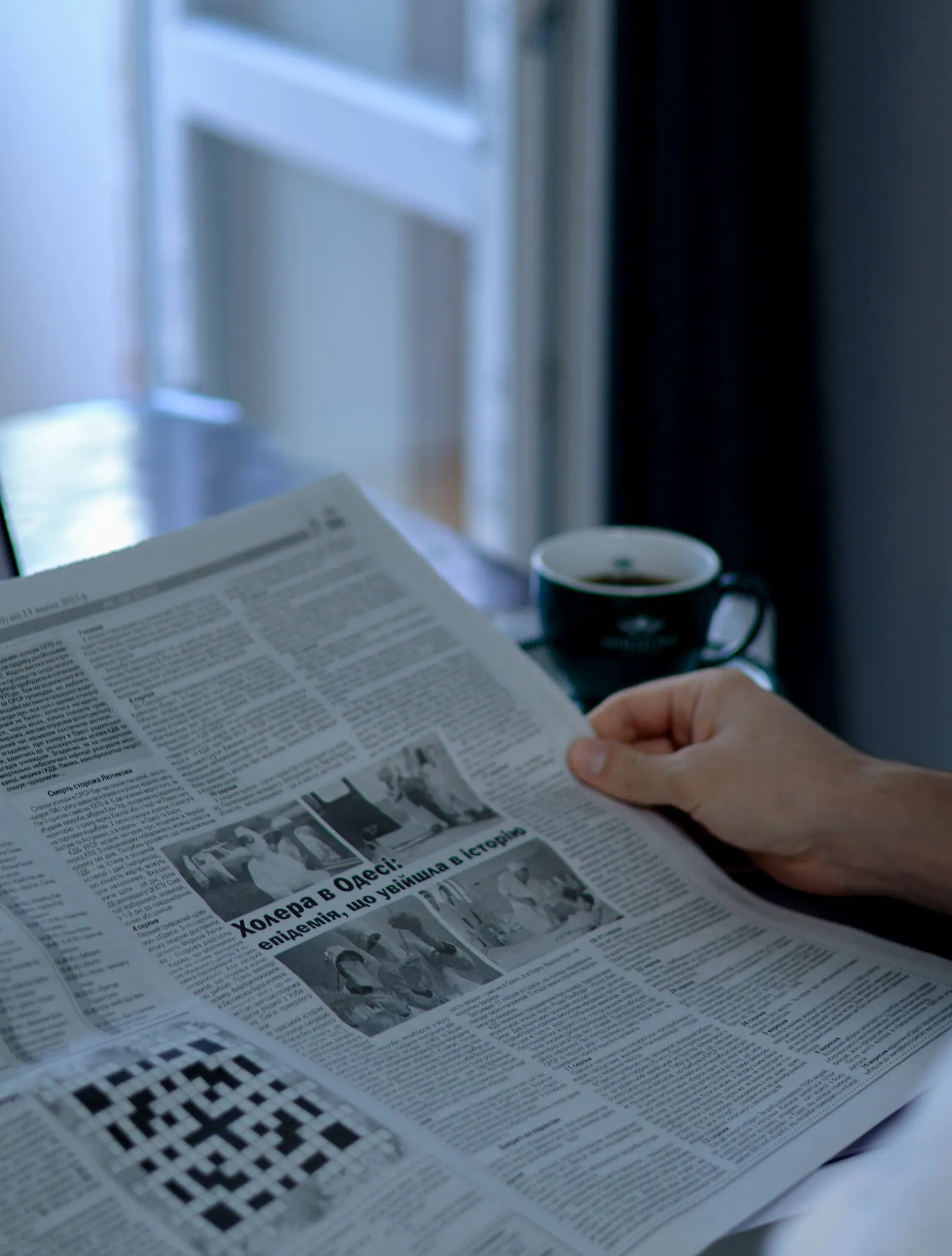 Person liest eine aufgeschlagene Tageszeitung, im Hintergrund eine Tasse Kaffee am Fenster