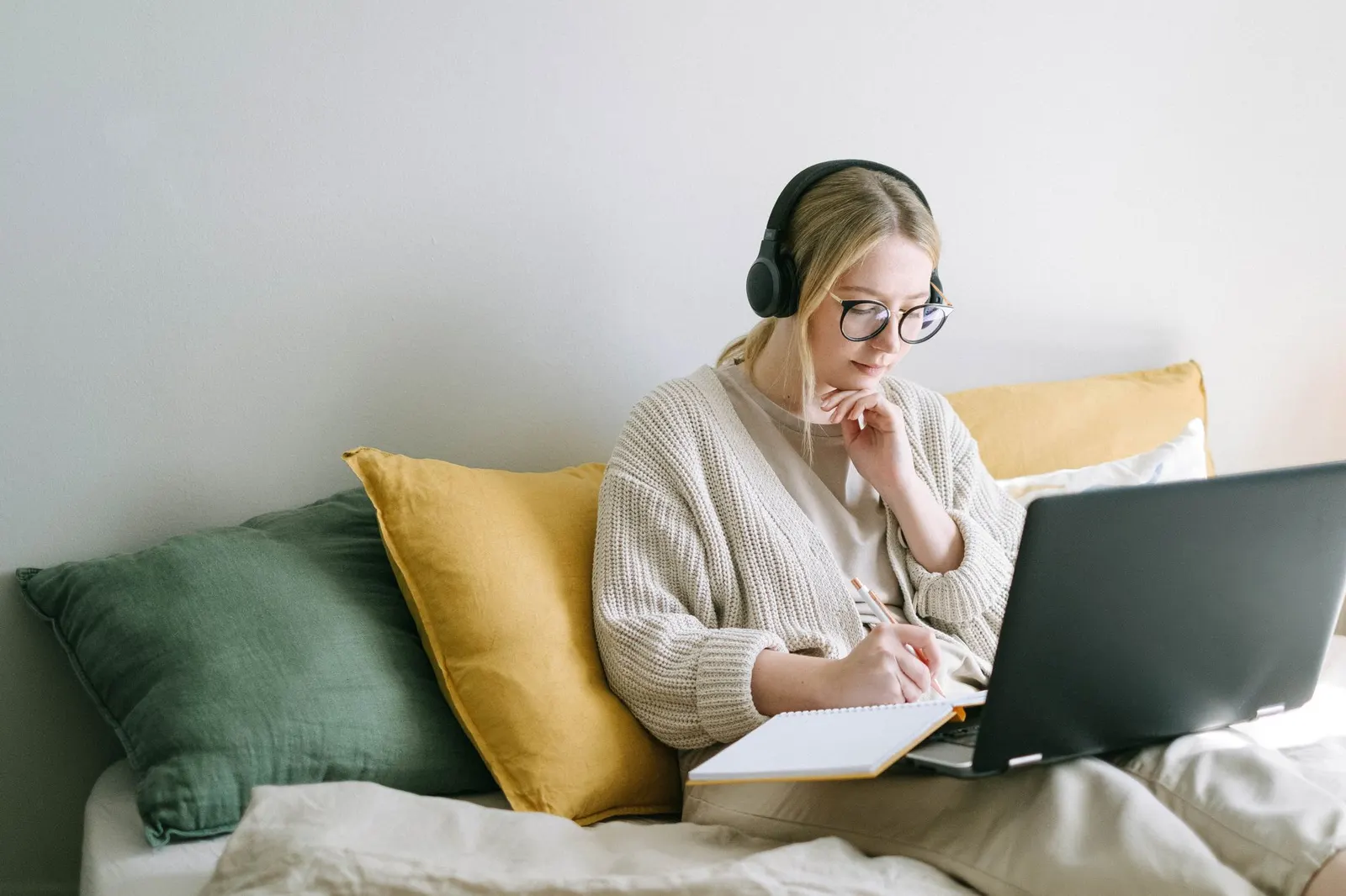 Frau mit Kopfhörern schreibt auf einem Sofa in ein Notizbuch und arbeitet am Laptop